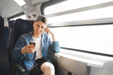Gülümseyen turistler seyahat tren ve kulaklık müzik dinler ve smartphone vasıl görünüyor. Mutlu yolcu treni pencerenin yakınında oturan ve Internet üzerinde smartphone kullanır