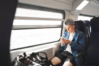 Bir tren seyahatlerinizde rahat kıyafetler giyen bir smartphone Internet'i kullanan ve kulaklık müzik dinler. Bir treni bir pencere yakınında oturan genç adam.