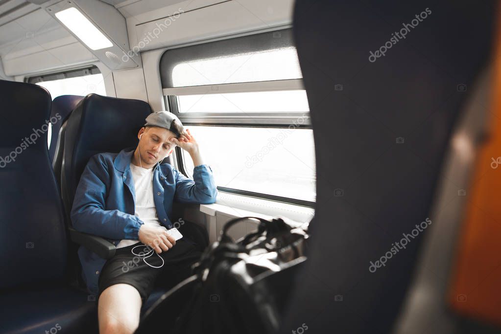 Young man sleeping in a train near the window with headphones and a smartphone in his hands. The sleepy guy goes by the train at the window.