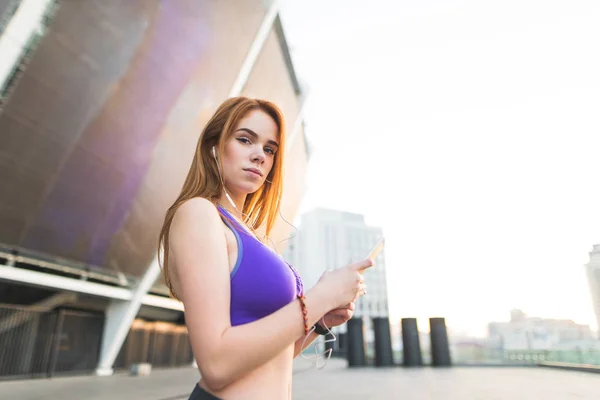 Attractive sports girl in the top, standing against the back of the stadium with a smartphone in his hands, listening to music in the headphones looking into the camera, resting after jogging.