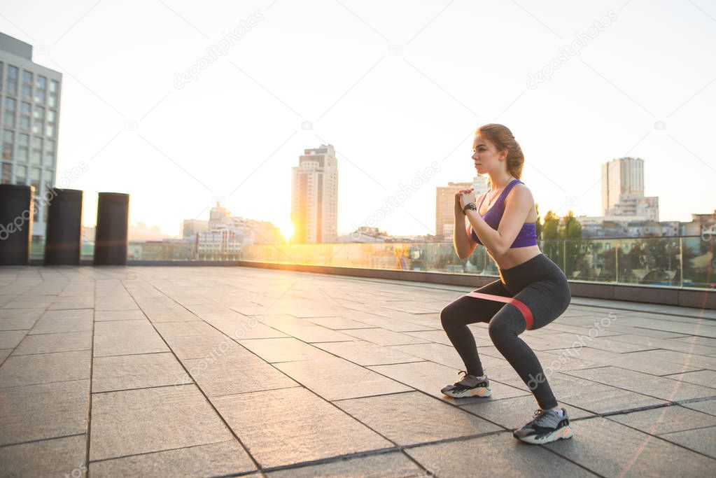 Portrait of a charming girl in sportswear doing training in the background of the sunset. Girl squats with a rubber band on the street. Sport in the morning.