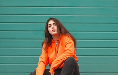 Yeşil duvar çerçevede bir parlak rahat elbiseli şık bir sokak kız portresi. Renkli bir arka plan üzerinde turuncu bir hoodie çekici bir kız sokak fotoğrafı