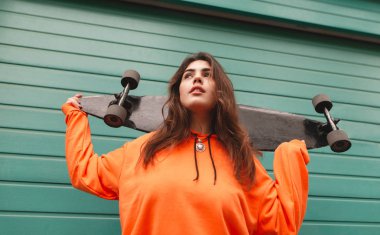 Onun elinde bir longboard ile rahat kıyafetler içinde çekici kız bir yeşil, Turkuaz duvar arka plan üzerinde duruyor. Renkli bir arka plan üzerinde bir kız patenci portresi. Sokak stili