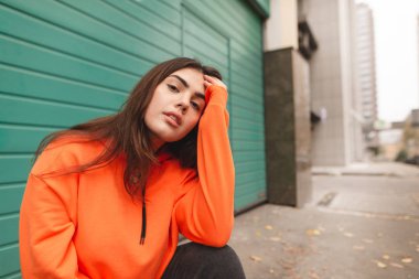 Sokakta oturan ve kameraya poz bir turuncu hoodie şık bir kız sokak portresi. Sokak giysileri sokakta oturan güzel kızı.