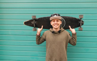 Gülümseyen genç bir kap ve kapüşonlu bir yeşil duvar arka plan üzerinde duruyor, omzunda bir longboard tutar, kameraya bakar ve sevinir. Şanslı patenci portresi. Boşaltmak
