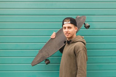 Casual giyim çekici genç adam turkuaz bir arka plan üzerinde elinde bir longboard ile standları ve kameraya benziyor. Boşaltmak Skate kavramı.