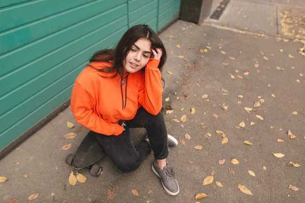 私服路上で座っていると笑顔の美少女スケーター ロングボードの上に座ってオレンジのパーカーでスタイリッシュな女の子のストリートの肖像 ストリート スタイルの十代の少女 — ストック写真