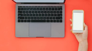 Kadının el tutar bir smartphone dizüstü turuncu zemin üzerine beyaz bir ekran ile yukarıdan görüntüleyin. Çalışma alanı ile bir smartphone el ve dizüstü bilgisayar, Flatlay.