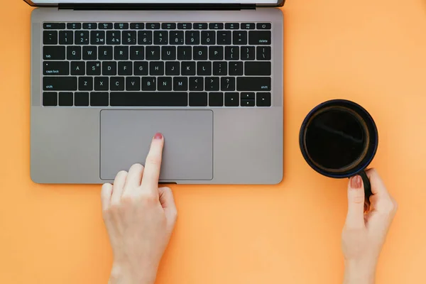 Woman uses a laptop on an orange background, a view from above. Hand holds a cup of coffee and uses a touchpad on the laptop. Flatlay