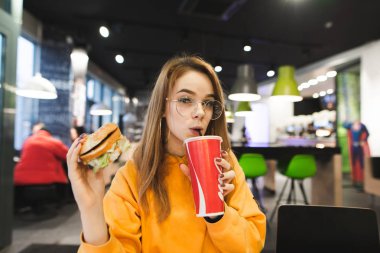 Onun elinde hamburger ile bir fast food restoranda oturan rahat elbiseli şık bir kız portresi kırmızı cam kola içer ve kameraya benziyor. Kız fast food yiyor
