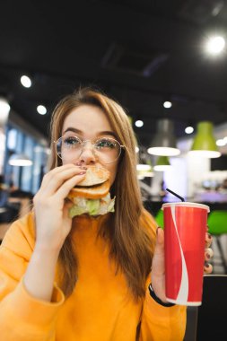 Lezzetli hamburger yemek yiyen ve içecek bir bardak onun elinde tutan eğlenceli, güzel bir kız portresi. Çekici kız bir ışık fast food restoran lokanta yiyor. Kız bir hamburger yiyor