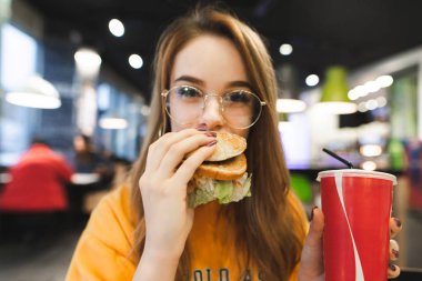 İştah açıcı bir burger ısırma, bir bardak kola onun elinde tutan, kameraya bakarak gözlüklü genç bir kız portresi kapatın. Kız bir fast food restoran ve içecek iştah açıcı bir hamburger yiyor
