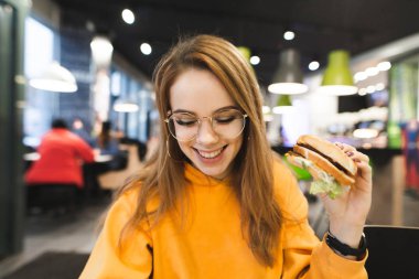 Olumlu bir parlak casual giyim ve gözlük güzel kız ellerini ve gülümsüyor hamburger ile bir fast food restoran oturur. Bir burger Restoran arka plan üzerinde gülümseyen kız