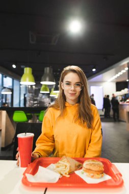 Kız bir kafede bir telefon için konuşmak, fast food yiyor, kamera ve gülümsüyor görünüyor. Bir fast food restoranında akşam yemeği portre sevimli öğrenci