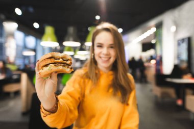 Parlak elbiseleri mutlu kız iştah açıcı bir büyük hamburger bir fast-food restoran ve gülümsüyor arka plan üzerinde tutar. Kız öğrenci burger el ile. Burger üzerinde odaklanmak.