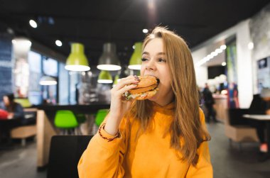 Turuncu kapüşonlu bir güzel kız portresi bir fast-food restoran çerçevede iştah açıcı bir hamburger yiyor. Kız burger ısırıkları ve zevk alır. Fast Food kavramı.