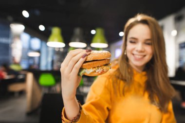 Pozitif güzel kız fast food restoran içinde oturur, lezzetli büyük hamburger elinde tutan ve gülümsüyor. Genç kız bir hamburger yiyor. Kızın elinde taze iştah açıcı hamburger odaklanmak.