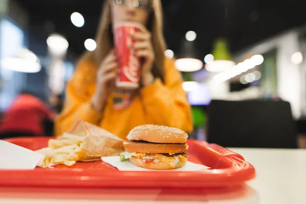 赤いグラスで飲み物を飲む女の子の背景にハンバーガーとフライド ポテトとトレイ ファーストフードのレストランで食事 ファーストフードのコンセプト — ストック写真