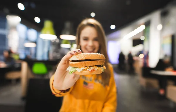 微笑的女孩坐在一家快餐店的桌旁 向镜头展示了一个开胃的大汉堡 美味的汉堡掌握在一个积极的女孩手中 专注于汉堡 — 图库照片