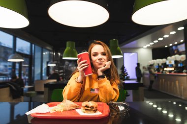 Tatlı kız bir fast-food restoran masada yemek tepsisiyle oturur, cola kırmızı bir bardak onun elinde tutan, kamera ve gülümsüyor görünüyor. Güzel kız öğrenci fast food yiyor