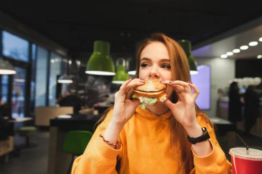 Parlak turuncu giysileri güzel kızı bir kafede iştah açıcı bir hamburger yiyor ve yana görünüyor. Lezzetli hamburger yiyor bir kız portresi