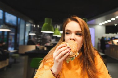 Tatlı kız büyük bir lezzetli burger iki el, ısırıkları ve içinde yakın çekim yan seyir tutar. Bir kızın yakın çekim portre restoran arka plan bir hamburger, hızlı yemek yiyor. Boşaltmak
