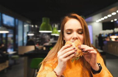Gözleri olan bir burger yemek zevk ile kapalı. Güzel bir kız ve bir burger fotoğrafı kapatın. Kız gözlerini kapatır ve arka planda restoranın lezzetli taze hamburger kokusu kokuyor.