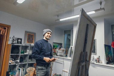 Genç sanatçı gülümseyen bir fırça ve palet bıçağı o boya resim ve gülümsüyor seyir stüdyo, elinde duruyor. Pozitif sanatçı rahat bir stüdyoda bir resmi boyar.
