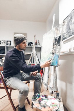 Yaratıcı adam bir stüdyoda oturan ve bir resim üzerinde çalışıyor. Profesyonel genç sanatçı yağlıboya resim kendi stüdyosunda boyar. Bir iş olarak boya.