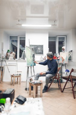 Ressam yağlıboya tablolar çizim rahat stüdyo ve inşaat üstünde bir tablo, bir sandalyede oturur. Sanatçı bir ev boyama stüdyoda çalışıyor. Bir hobi ve iş olarak boya.