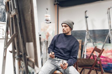 Genç profesyonel sanatçı bir şövale bir tuval ile yakın bir sandalyede oturur, resme bak bir fırça elinde ve şirin bir bakışla almıştır. Resmin üzerine çalışan ressam portresi.