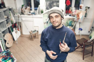 Ellerinde fırçalar ile kendi atölyesini ayakta, kamera seyir ve gülümseyen bir genç profesyonel sanatçı portresi. Ressam resim sergisi, Sephalar, ana bilgisayarlar ve paletleri ile bir stüdyo arka plan üzerinde.