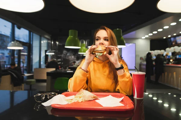 Girl bites the burger and gets pleasure. Fast Food Concept. Portrait of a beautiful girl in a orange hoodie bites an appetizing burger against the background of a fast-food restaurant.