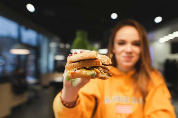 Attractive girl in orange clothes holding a big tasty burger, focus on burgers. Young girl holding in female hands fast food burger, american unhealthy calories meal on background.