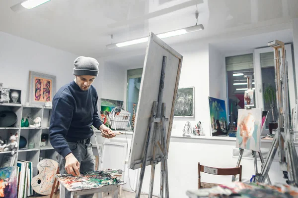 Young, positive artist works, paints a picture of oil paints in a cozy studio. Artist with a canvas on an easel places an oil paint on a palette. Process of creating a picture. Copyspace
