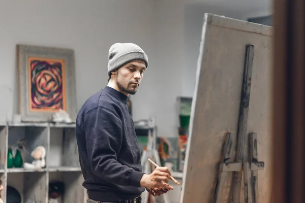 忙碌的男人艺术家 手里拿着一把刷子 在一个舒适的工作室里拿起一幅油画 才华横溢的年轻画家在画布上画了一幅画 绘画理念 作为一种爱好工作 — 图库照片
