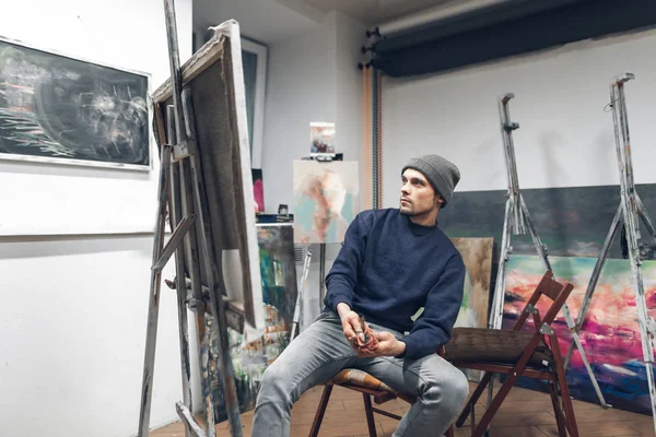 一个专业艺术家的肖像在一个舒适的家庭工作室的内部 画家坐在画架附近的椅子上 上面有主人和调色板 看着照片的严肃 绘画理念 — 图库照片