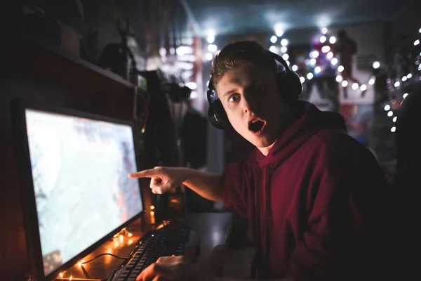 Surprised gamer,sitting at a desk near a computer, shows a finger on the screen and looks at the camera.Amazed teenager plays video games at home in his computer and looks emotionally at the camera