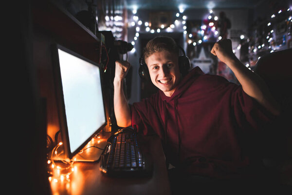 Портрет эмоционального подростка, сидящего дома за компьютером и радующегося победе с поднятыми руками. Радостный подросток играет в видеоигры дома на компьютере, смотрит в камеру и улыбается
