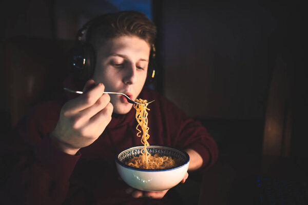 Close portrait of a young gamer eating spaghetti soup at a computer at night. Young man dinner at night with a noodle soup