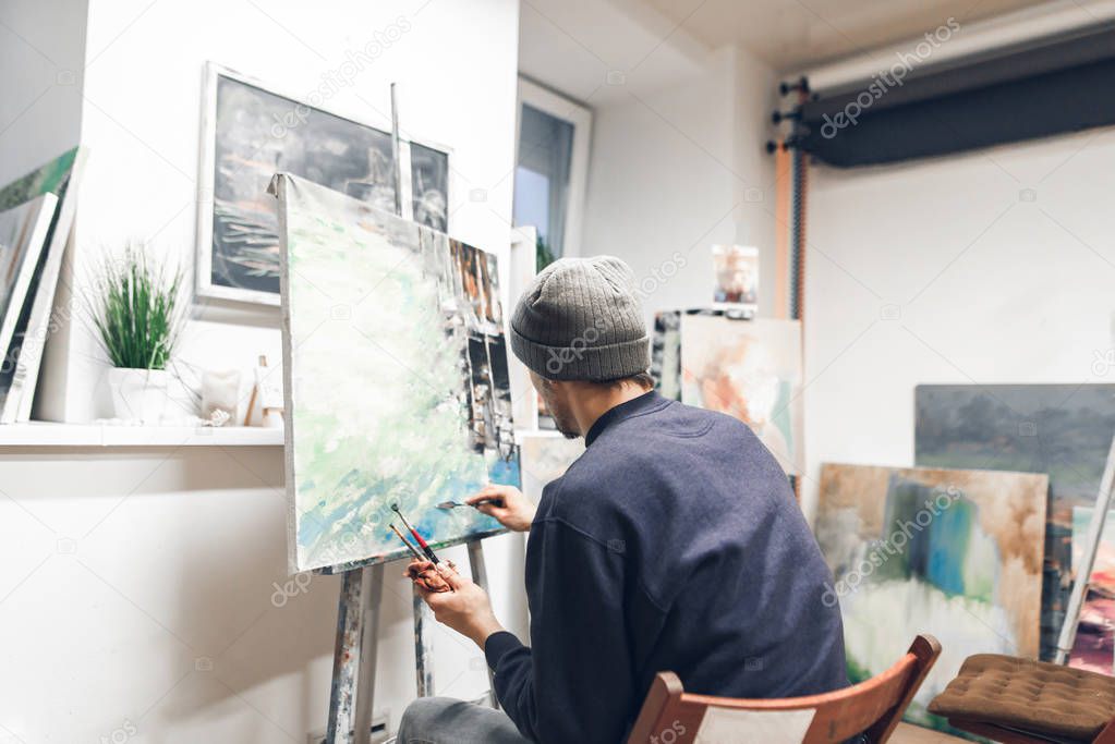 Back of a creative man painting a picture of oil in a home studio. Painter sits at home with brushes and a knife palette in his hands and creates a picture. Painting Concept. Hobby painting.