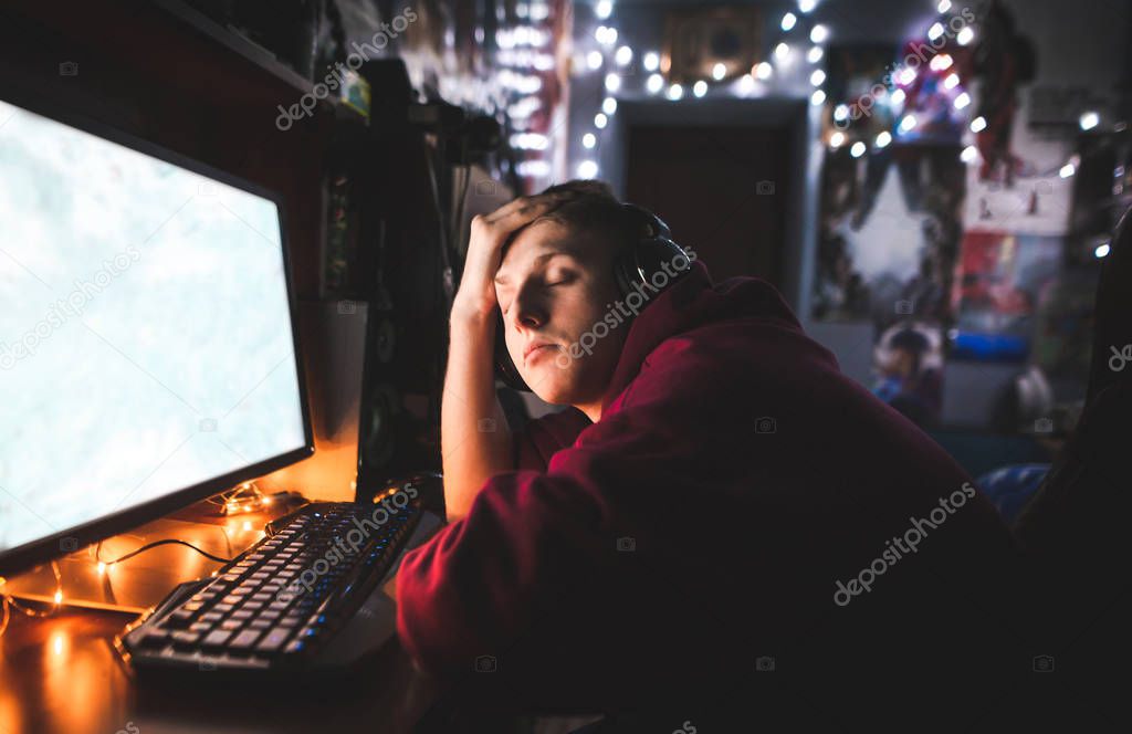 Portrait of a young gamer with headphones, plays games at night on the computer, and falls asleep on the table. Young gamer plays at home games at night and sleeps.