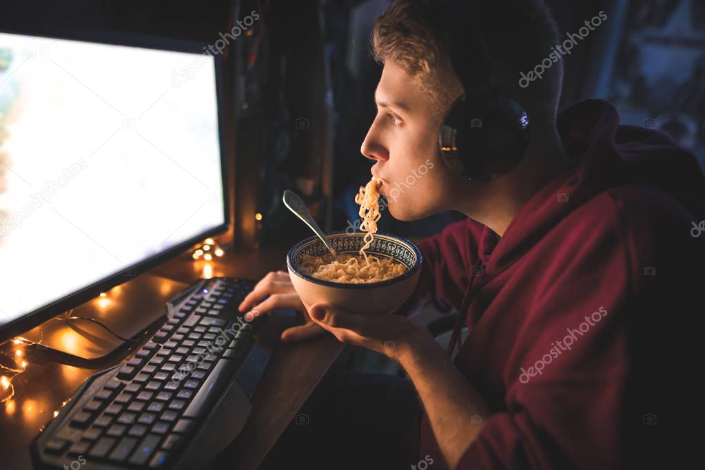 Portrait of a young man eating a noodle soup and enjoying a computer at night at home. A teen plays video games on a computer and eats fast food. Supper at the computer at night.