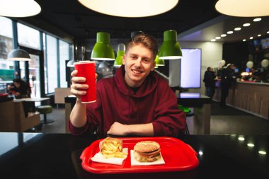 Genç adam gülümseyen bir bardak içki elinde tutan bir lokanta tepsisi ile masada oturuyor. Kamera ve gülümsüyor arıyorum. Pozitif öğrenci fast food yiyor