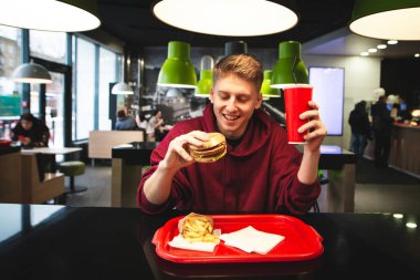 Bir fast food restoran masada oturuyor, bir burger holding ve cam içme, zararlı yiyecek arayan ve gülümseyen pozitif genç adam. Restoran masada lezzetli yemekleri ile mutlu öğrenci.