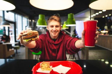 Mutlu genç adam bir burger tutar ve elinde içecekler, kamera gösterir. Burger ve kırmızı içecek camına odaklan. Olumlu bir genç adam elinde zararlı lezzetli yemek.