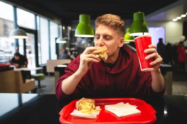 Aç öğrenci bir hamburger yiyor ve bir içki bir fast-food restoran önünde tutar. Genç adam lokanta hamburger yeme ve zevk bir restoran masada yemek yer. Fast Food kavramı.
