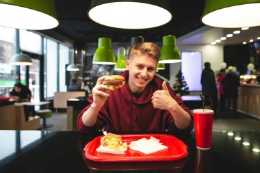 Bir fast-food restoran bir masada oturan bir tepsi ve onun elinde hamburger ile çekici genç adam bir başparmak yukarıya, gülümsüyor gösterir ve kameraya benziyor. Gibi bir öğrenci bir burger el ile gösterir