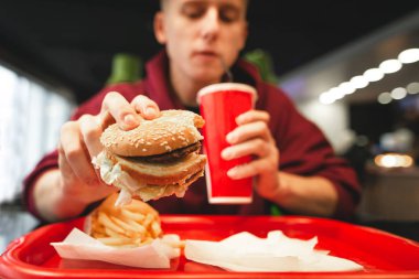 Genç adam, bir fast food restoran yemekleri lokanta. Adam bir burger kırmızı cam dışında bir kola içme, elinde tutar. Patates kızartması ve lokanta tepsi. Fast Food kavramı. Genç fast food yiyor