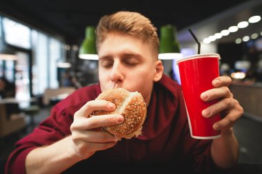 Bir bardak içki elinde tutan komik genç adam gözleri kapalı, lezzetli bir burger yemek zevk ile. Yakın çekim portre bir adam yeme fast food. Öğrenci bir burger yakın çekim fotoğraf yiyor